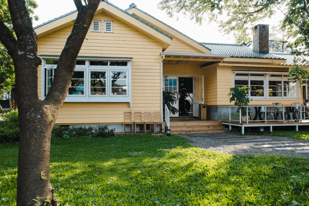 Image shows the exterior of a home with yellow siding and white windows with a tree in front of it