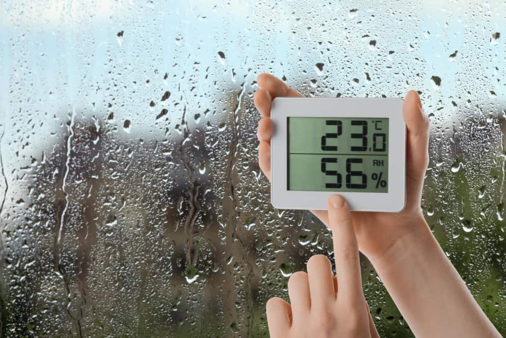 Image depicts a person holding a digital hygrometer in front of a condensation-covered window. The hygrometer reads 23 degrees Celsius and 56% humidity.