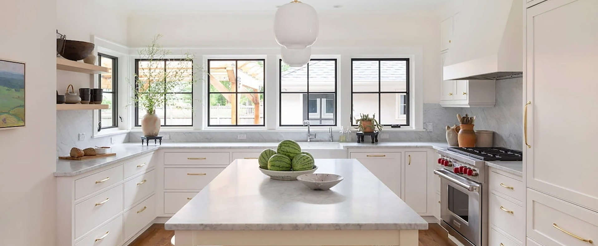 A bright and modern-style kitchen with all white cabinetry and marble countertops. The black wood-framed Pella windows line the far wall.