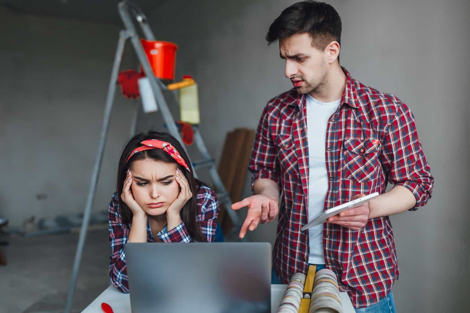 Image shows a man and woman looking at a laptop in a room being remodeled. The woman's hands are on her temples and the man appears upset. 