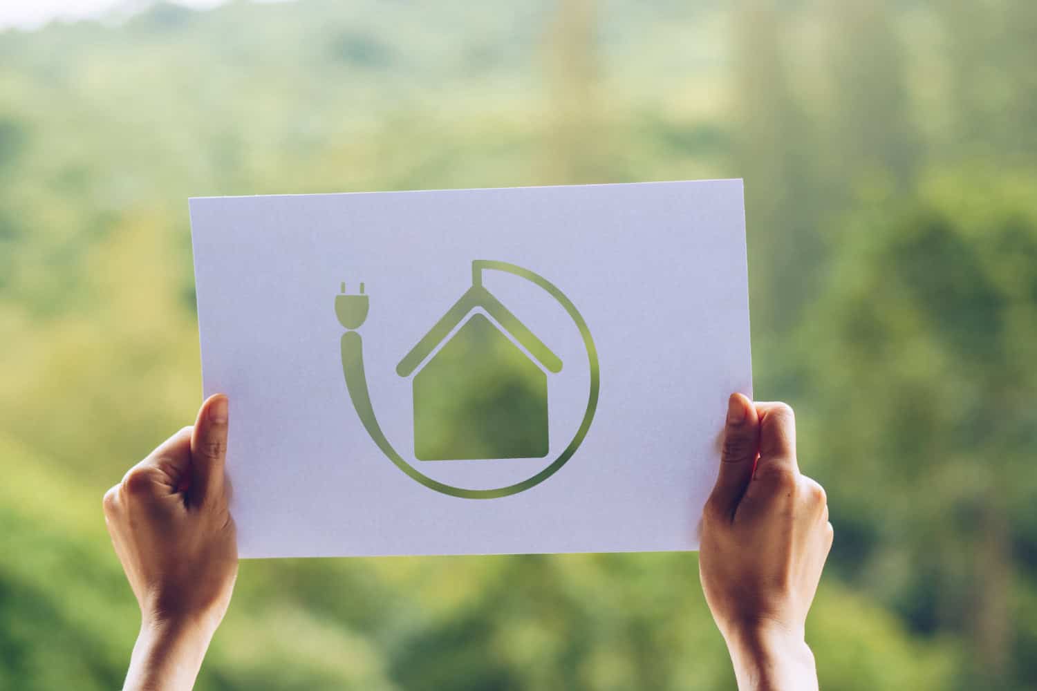 Image shows a woman's hands holding a sheet of paper up to a window. The paper has a translucent green logo of an energy efficient home in the center.