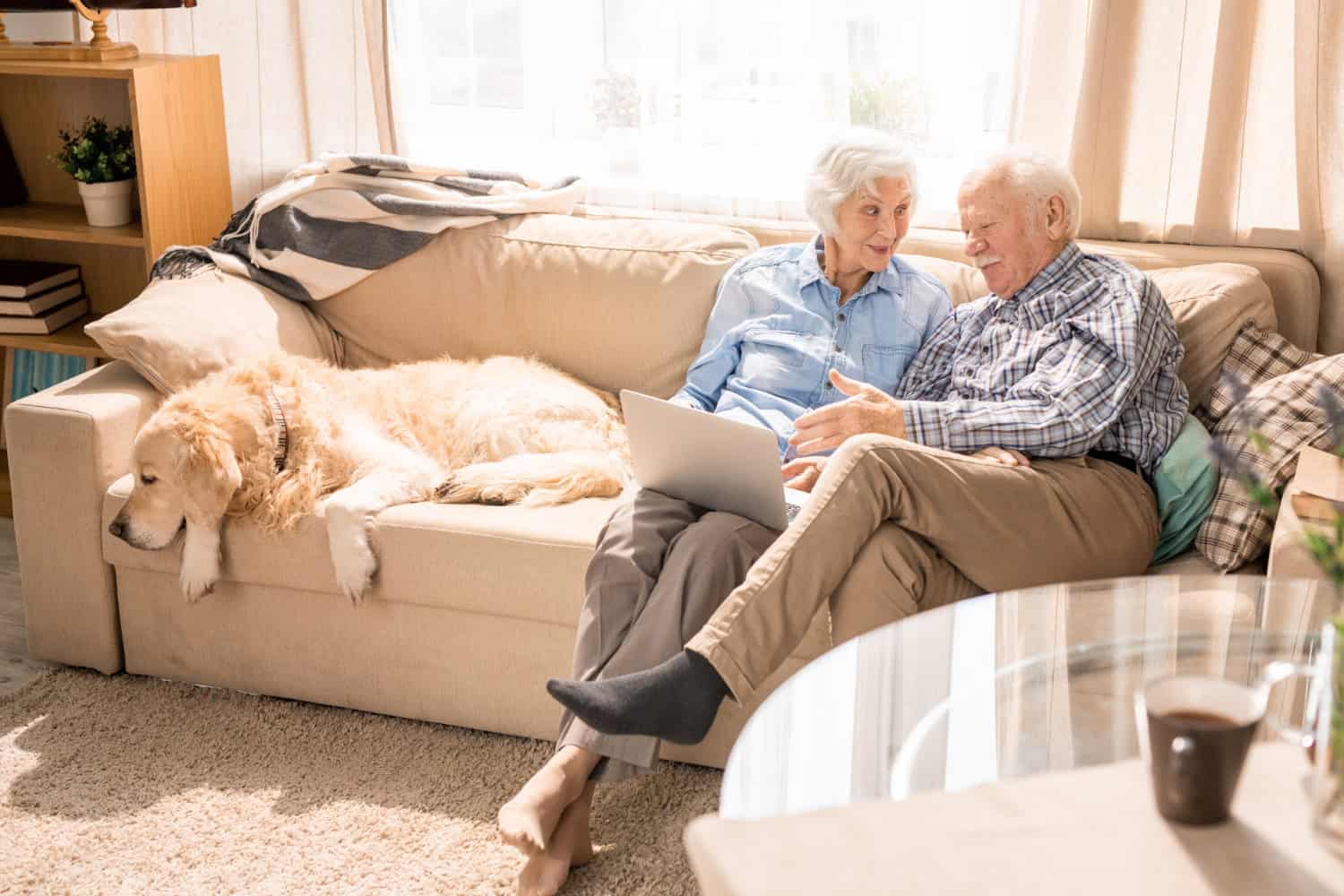 Image shows an older couple seated on a couch in their home looking at a laptop computer. Their large, cream colored dog lays next to them on the couch. 