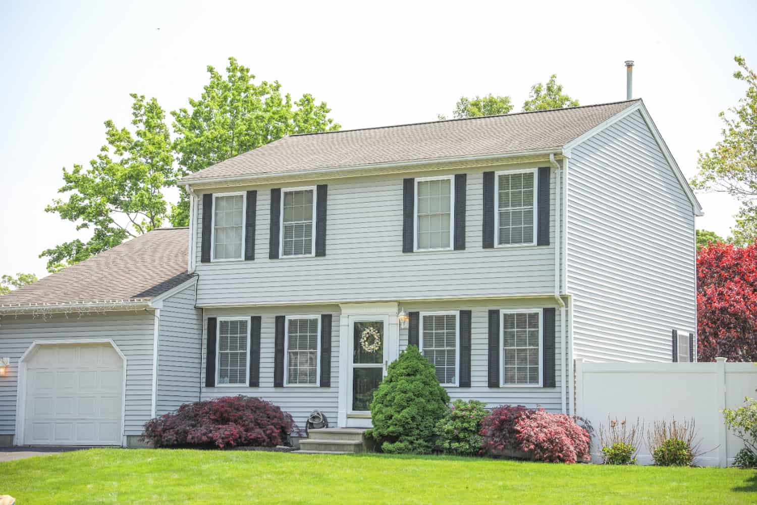 A classic two-story home with white siding, windows lining the front of a home, and a manicured lawn, pictured on a clear, sunny day.