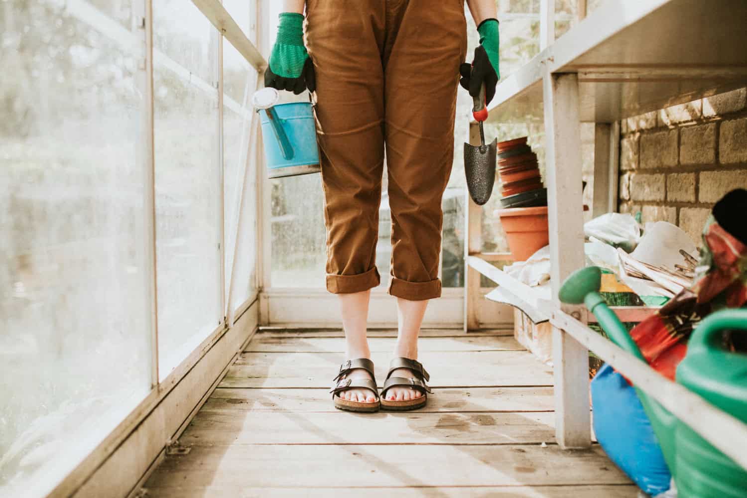 A woman pictured from the waist down is standing in a workshop, holding a can of paint and a gardening shovel. She's wearing capri pants, sandals, and work gloves.